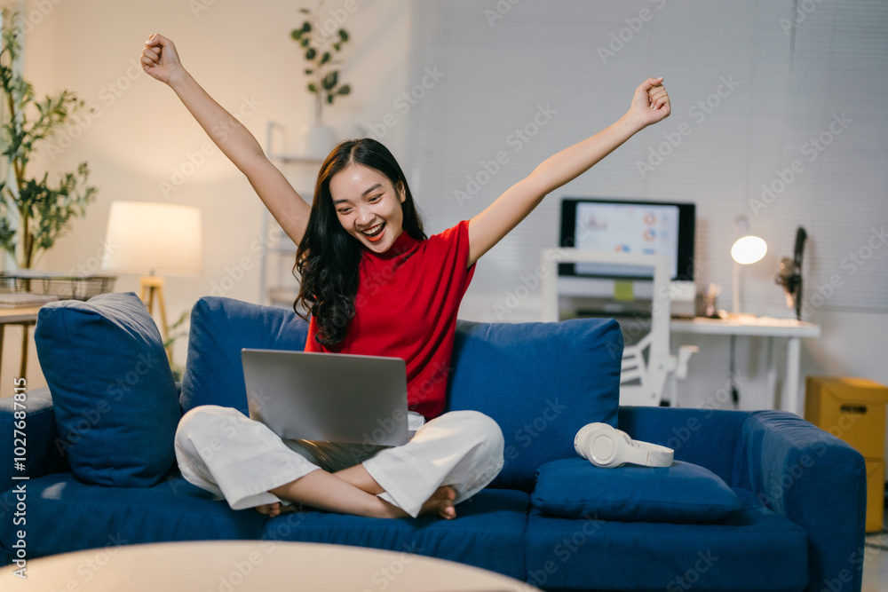 Young asian woman is sitting on her sofa with her laptop, she is raising her arms in the air with a big smile on her face, celebrating a success Young asian woman is sitting on her sofa with her laptop, she is raising her arms in the air with a big smile on her face, celebrating a success