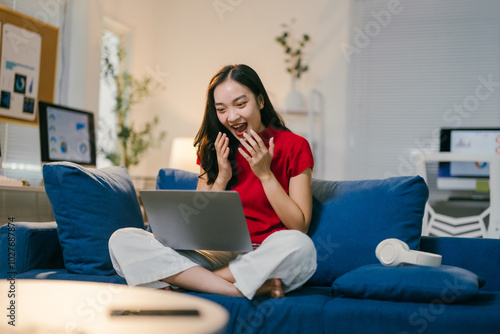 Young woman is sitting on her couch working from home. She is looking at her laptop computer and reacting with excitement to something she is seeing