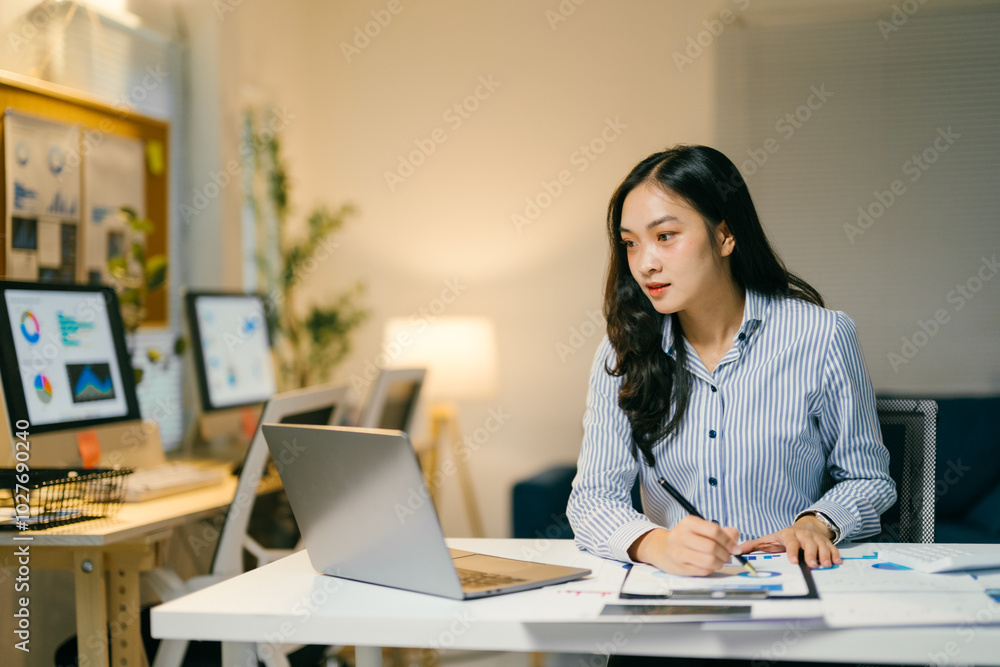 Young professional is reviewing charts and data on her laptop, taking notes on a notepad in her brightly lit workspace Young professional is reviewing charts and data on her laptop, taking notes on a notepad in her brightly lit workspace
