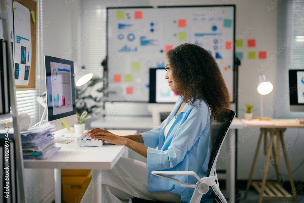 Obraz premium Young professional is analyzing data on her computer in a bright, modern office. She is surrounded by charts and graphs, indicating her focus on business or finance