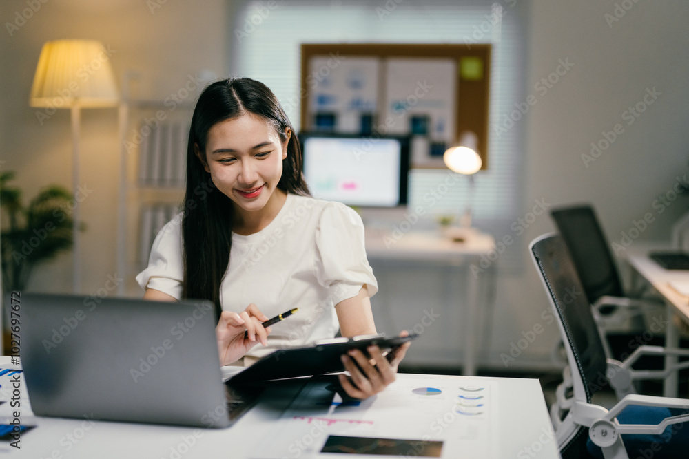 Young businesswoman smiles while analyzing documents and working on her laptop in her office, exuding confidence and cheerfulness Young businesswoman smiles while analyzing documents and working on her laptop in her office, exuding confidence and cheerfulness