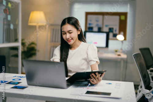 Businesswoman is reviewing financial data on her laptop and taking notes on a clipboard while sitting at her desk in a modern office