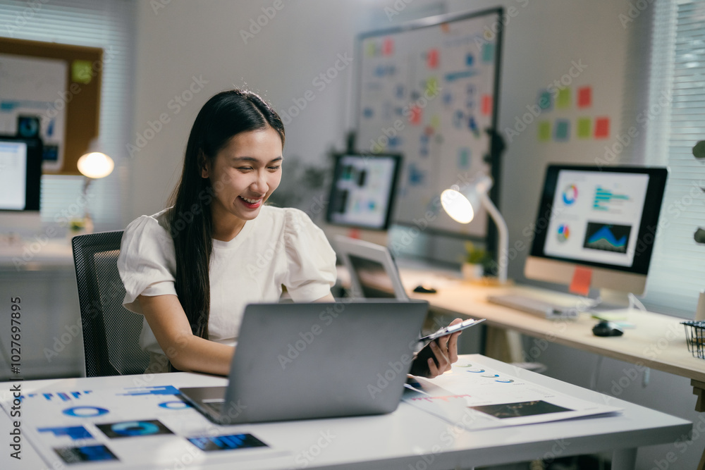 Young professional is analyzing charts and graphs on her laptop while holding a digital tablet in her other hand, smiling at the positive results Young professional is analyzing charts and graphs on her laptop while holding a digital tablet in her other hand, smiling at the positive results