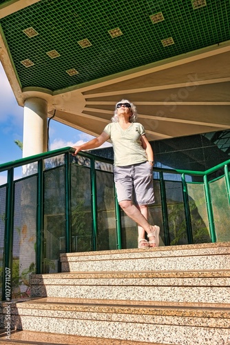 An elderly woman in black sunglasses stands on the stairs of a modern building and looks up.