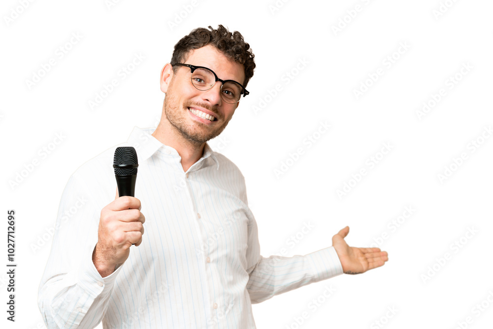 Brazilian man picking up a microphone over isolated chroma key background extending hands to the side for inviting to come Brazilian man picking up a microphone over isolated chroma key background extending hands to the side for inviting to come