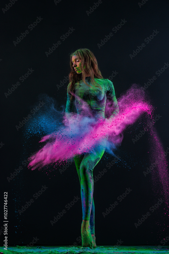Obraz premium girl dancer in black suit dancing with explosions of colored paints holi on dark background