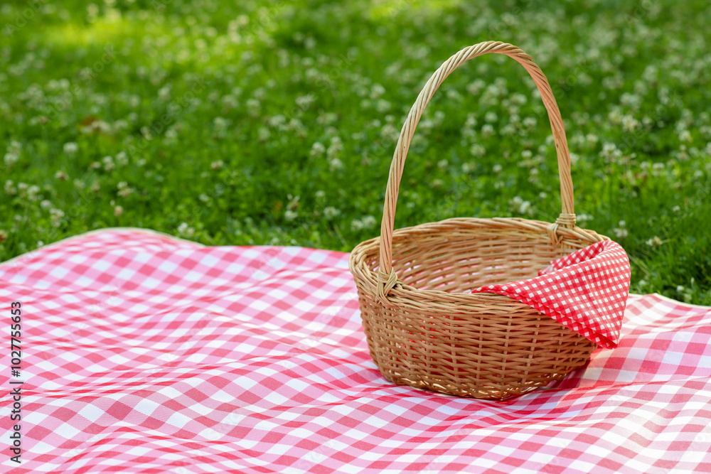 One picnic wicker basket with checkered napkin and blanket on green grass. Space for text One picnic wicker basket with checkered napkin and blanket on green grass. Space for text