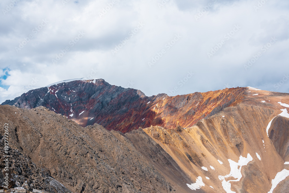 Colorful layered landscape with sunlit sharp rocky mountain ridge of vivid colors under cloudy sky. Golden rocks and red sheer crags in sunlight. Shadows of clouds on unusual multicolored mountains. Colorful layered landscape with sunlit sharp rocky mountain ridge of vivid colors under cloudy sky. Golden rocks and red sheer crags in sunlight. Shadows of clouds on unusual multicolored mountains.