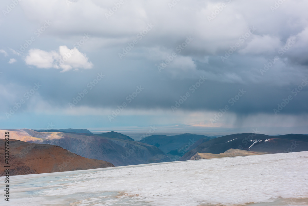 Fototapeta premium Dramatic alpine top view from flat sloping glacier on precipice edge to deep gorge between colorful sunlit cliffs and mountain range silhouette in rain under lead gray cloudy sky. Rainy grey clouds.