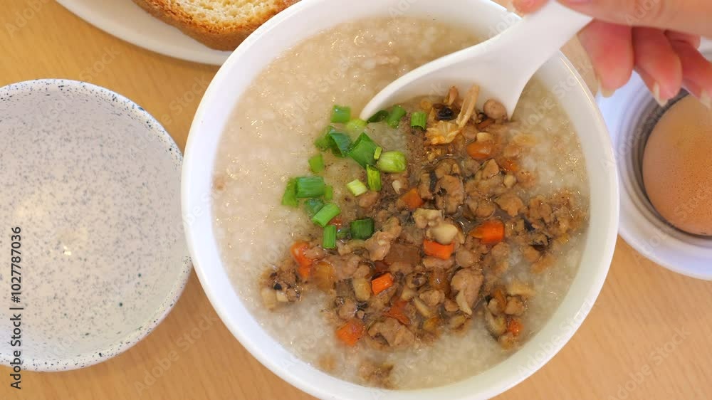 Close-up of bowl of freshly made congee, garnished with cooked minced pork, chopped green onions, and carrots, with ceramic spoon ready to serve. Comfort Food and Traditional Cuisine. Close-up of bowl of freshly made congee, garnished with cooked minced pork, chopped green onions, and carrots, with ceramic spoon ready to serve. Comfort Food and Traditional Cuisine.
