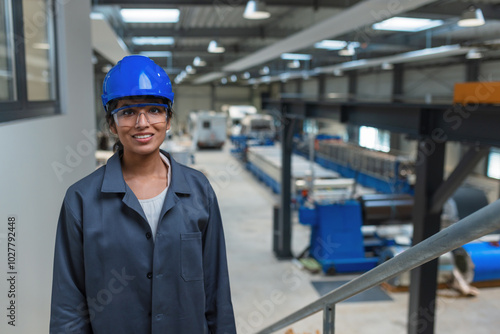 Confident young engineer, an Indian woman, in a blue helmet and gray work coat at a factory, portrait shot.