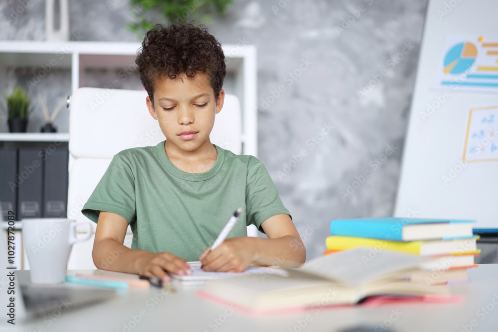 Portrait of a cute African American kid student sitting at the table working on his hometask, making notes at the copybook Portrait of a cute African American kid student sitting at the table working on his hometask, making notes at the copybook