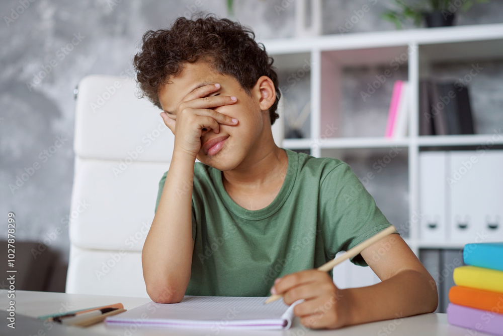 Close up portrait of an African American kid student covering his face with hand feeling tired and sad unwilling to continue working on his home assignment Close up portrait of an African American kid student covering his face with hand feeling tired and sad unwilling to continue working on his home assignment