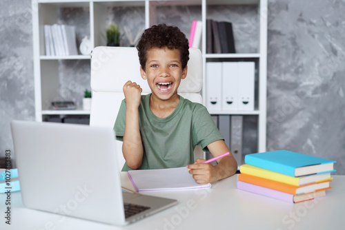 Happy smiling African Americans schoolboy sitting at the tableand making winner's gesture clenching his fist while working on his hometask photo