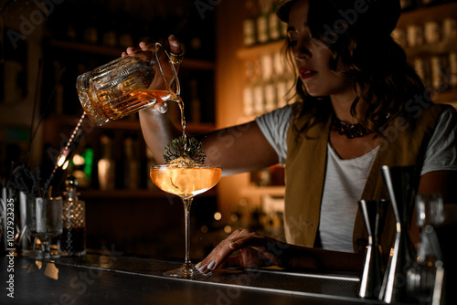 Female bartender pours a cocktail from a mixing glass into a tall stemmed glass with an ice cube