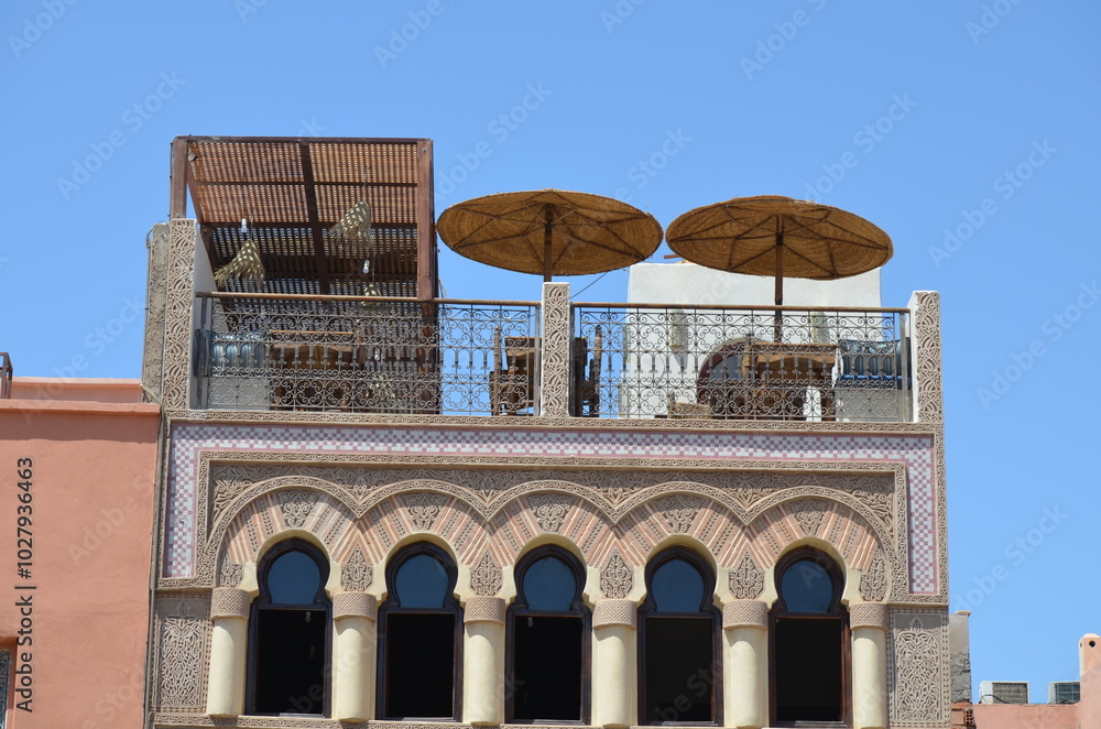 Obraz premium Traditional Moroccan terrace with decorative arches, ornate ironwork, and woven parasols in Marrakech, under clear blue sky 