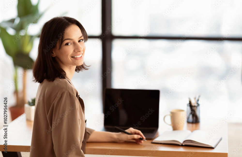 Online Education. Smiling young teacher holding pen looking at camera, sitting at desk. Laptop mock up, empty space Online Education. Smiling young teacher holding pen looking at camera, sitting at desk. Laptop mock up, empty space