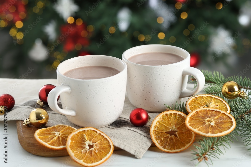 Tasty hot cocoa drinks in cups, fir branches, baubles and dry orange slices on white table against blurred lights, closeup Tasty hot cocoa drinks in cups, fir branches, baubles and dry orange slices on white table against blurred lights, closeup