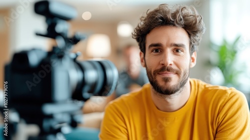 A confident man in a yellow shirt is posed with a camera setup, amidst a soft-focused, modern environment, suggesting creativity and professionalism.