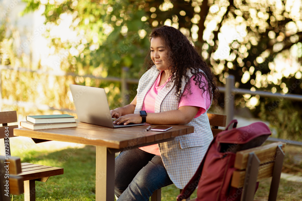 A student is engaged with a laptop outdoors, surrounded by greenery, enjoying the sun while studying. Books are stacked beside her, and a backpack rests on the chair. A student is engaged with a laptop outdoors, surrounded by greenery, enjoying the sun while studying. Books are stacked beside her, and a backpack rests on the chair.