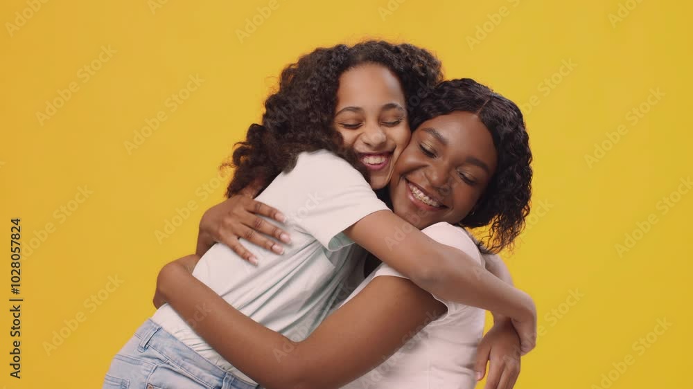 A young black girl with curly black hair is hugging an adult woman who also has curly black hair. They are both wearing white t-shirts and smiling. The background is a bright yellow. A young black girl with curly black hair is hugging an adult woman who also has curly black hair. They are both wearing white t-shirts and smiling. The background is a bright yellow.