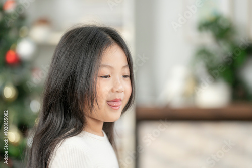 A young girl with long, dark hair smiles gently in a softly lit room, surrounded by a warm and festive environment. The scene conveys a sense of calm, comfort, and innocence.