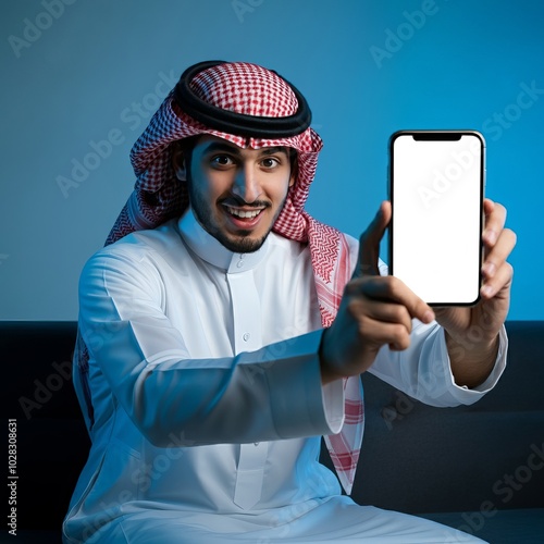 Young Arab muslim man in traditional clothes showing mobile phone isolated on home and sitting on sofa with blue colors.
