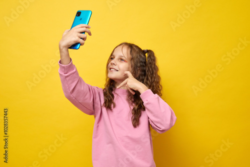 Little girl recording video or making selfie for social media with mobile phone excited to share joyful mood with her online audience showing heart gesture isolated over yellow background photo