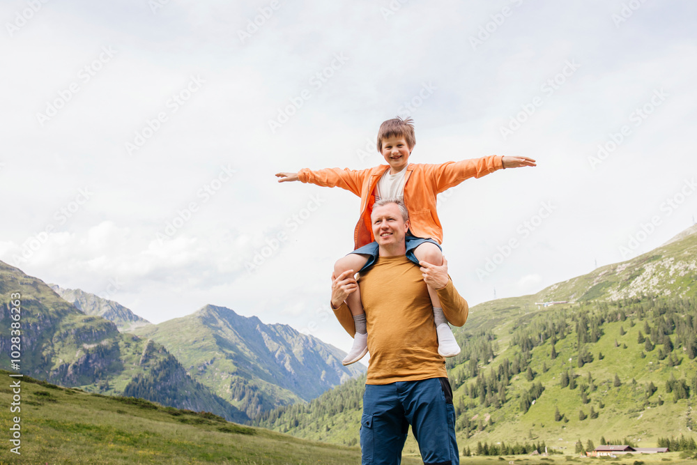 Happy boy with arms outstretched sitting on father's shoulder at meadow Happy boy with arms outstretched sitting on father's shoulder at meadow