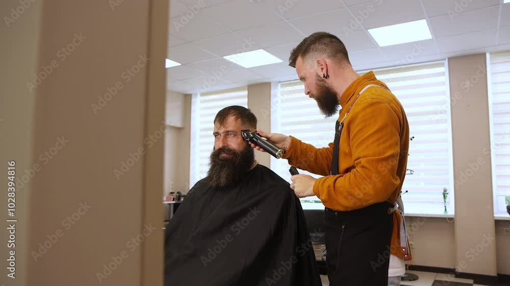 Stylish handsome barber in orange clothes and apron doing haircut to man and trimming beard in barber shop Stylish handsome barber in orange clothes and apron doing haircut to man and trimming beard in barber shop