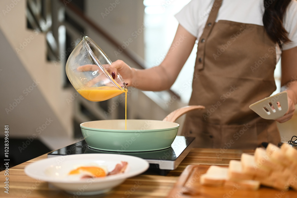 Young woman in apron pouring a beaten eggs from a glass bowl into a frying pan Young woman in apron pouring a beaten eggs from a glass bowl into a frying pan