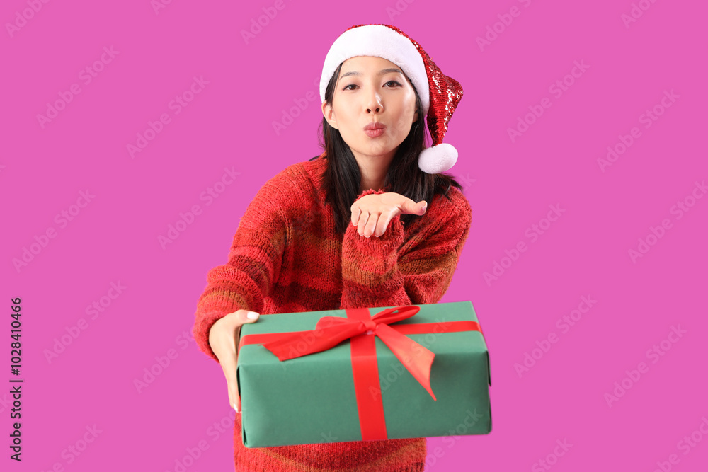Young beautiful Asian woman in Santa hat with gift box blowing kiss on purple background Young beautiful Asian woman in Santa hat with gift box blowing kiss on purple background