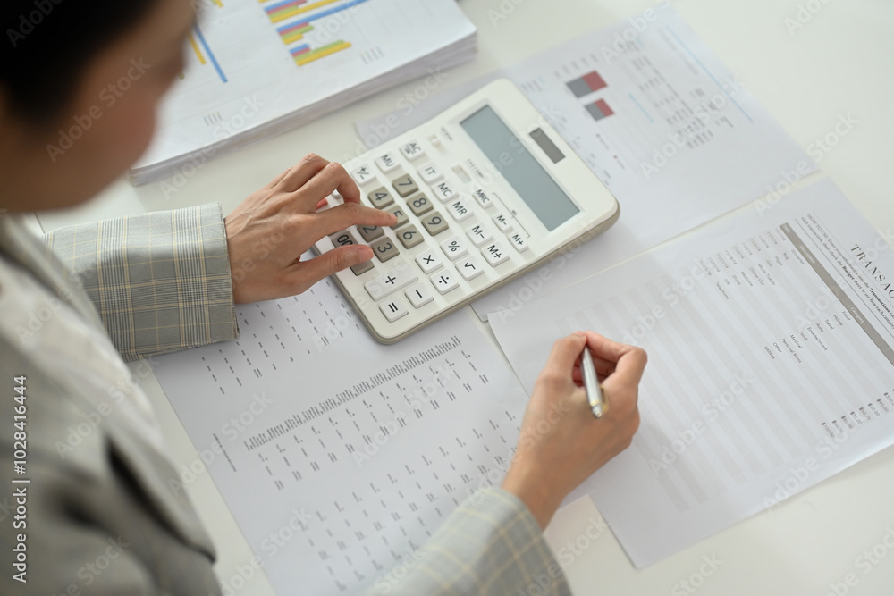 Female accountant using calculator and reviewing reports with charts and transaction details Female accountant using calculator and reviewing reports with charts and transaction details
