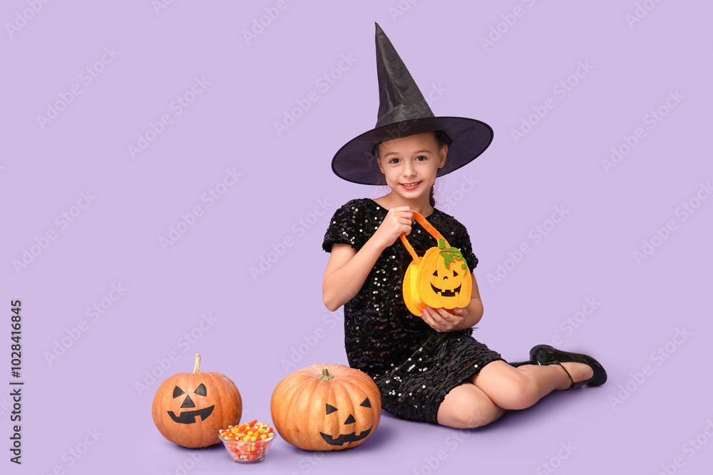 Fototapeta premium Cute little girl dressed for Halloween as witch with pumpkins, bag and candies sitting on purple background