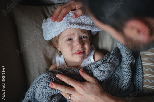 Father taking care of sick son with fever and cold, holdig cold cloth to his forehead. Boy lying on sofa, feeling tired. Top view.