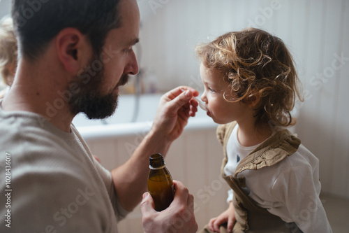 Sick girl with fever and cold at home. Father giving his daughter medicine on spoon, cough syrup or fever medicine.