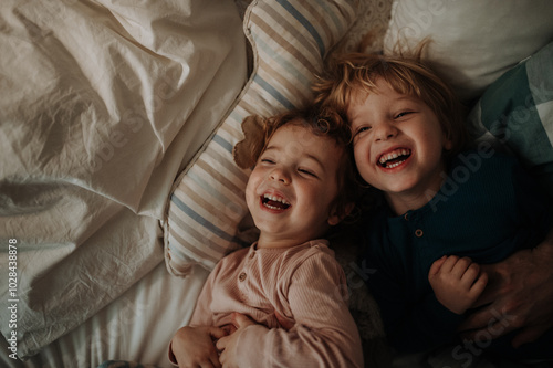 Schilderij op canvas Candid portrait of siblings lying together in bed, looking at the camera and laughing