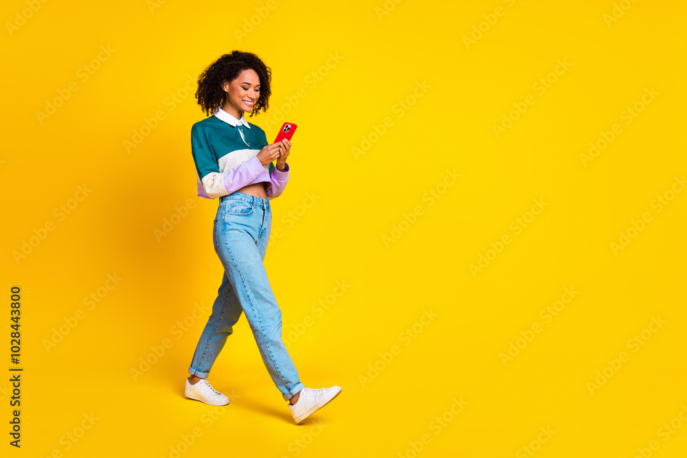 Full length photo of optimistic girl wear striped shirt look at smartphone go empty space isolated on vivid yellow color background Full length photo of optimistic girl wear striped shirt look at smartphone go empty space isolated on vivid yellow color background