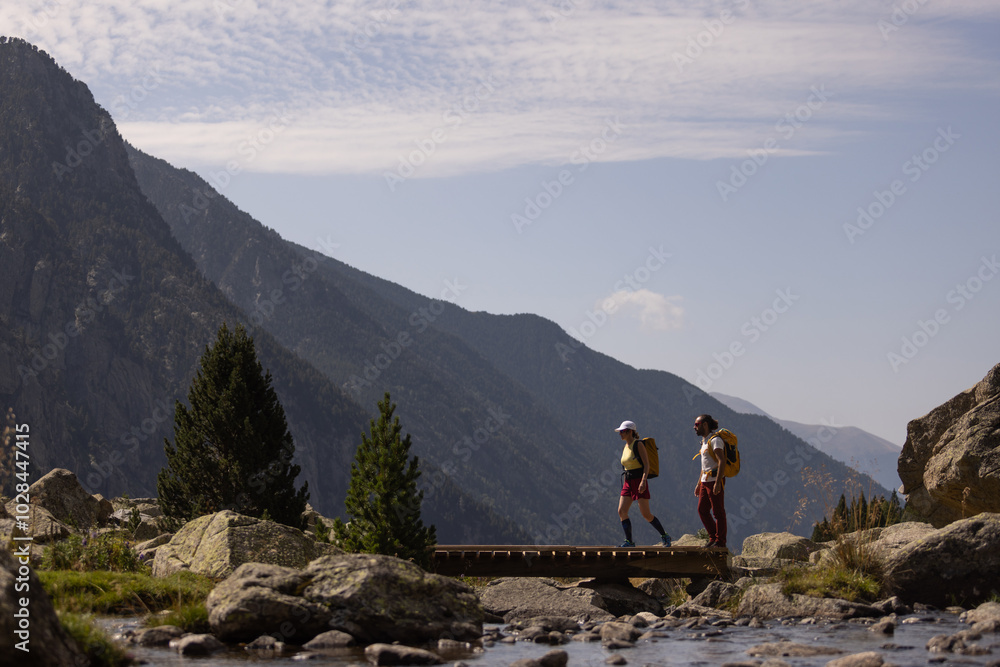 Fototapeta premium Two people are walking across a bridge over a river. The mountains in the background create a sense of awe and wonder. The scene is peaceful and serene, with the sound of the water