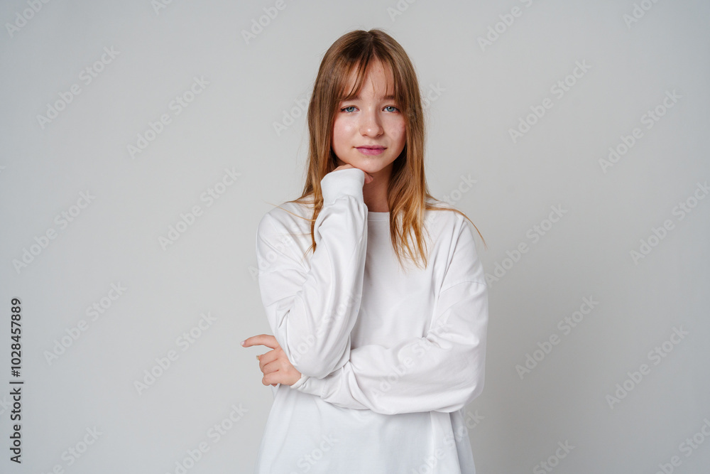 Young woman in a white long-sleeve shirt thoughtfully poses with a hand on her chin in a minimalistic setting Young woman in a white long-sleeve shirt thoughtfully poses with a hand on her chin in a minimalistic setting