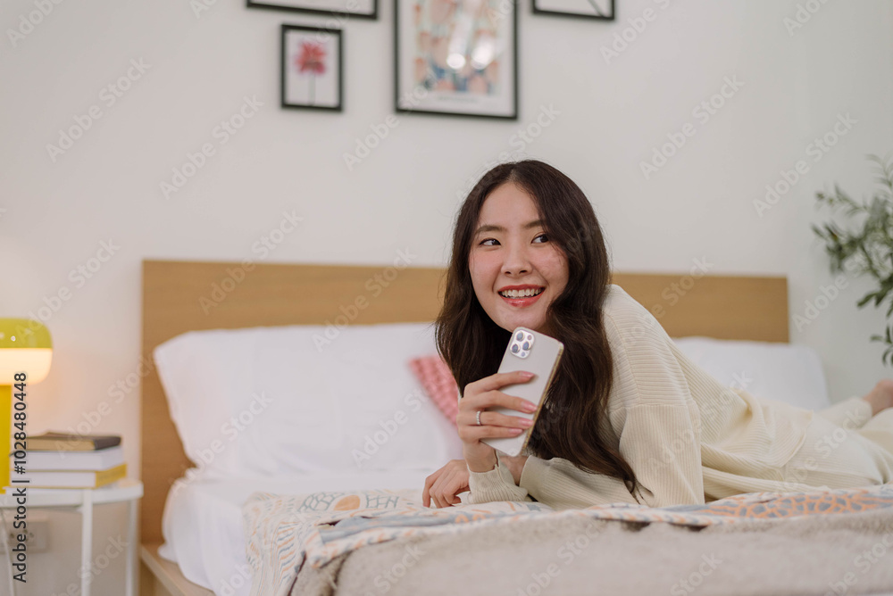 Asian woman in cozy attire smiling while using smartphone after wake-up on a bed in bedroom with a morning warm ambiance. Lifestyle and daily activities. Asian woman in cozy attire smiling while using smartphone after wake-up on a bed in bedroom with a morning warm ambiance. Lifestyle and daily activities.