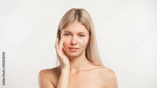 Young blonde woman posing at camera applying face cream on cheek skin and smiling, isolated on white studio background. Beauty skin care concept