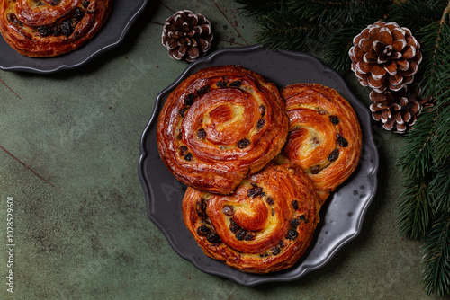 Pain aux raisins, also called escargot or pain russe, is a spiral pastry with custard cream and raisin on a green decorated table with christmas pastry, tree branch and pine. Green table. photo