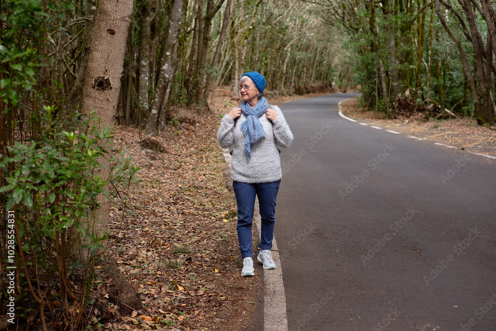Front view of elderly caucasian woman enjoying freedom in outdoors walking in a mountain road. Elderly active woman traveling in a forest holding backpack enjoying nature and vacation Front view of elderly caucasian woman enjoying freedom in outdoors walking in a mountain road. Elderly active woman traveling in a forest holding backpack enjoying nature and vacation