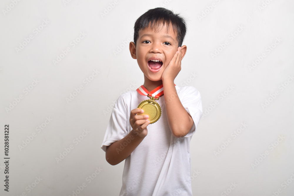 Little asian boy with a surprised expression holding a gold medal for the first place isolated on a white background. The boy celebrating his success Little asian boy with a surprised expression holding a gold medal for the first place isolated on a white background. The boy celebrating his success