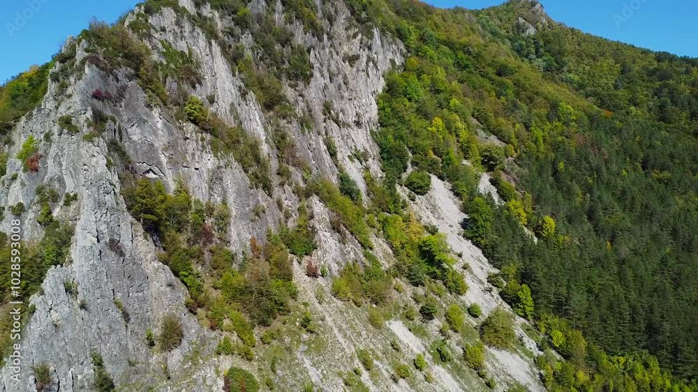 Drone view of a rocky mountain covered by vegetation, revealing houses below, on a sunny day Drone view of a rocky mountain covered by vegetation, revealing houses below, on a sunny day