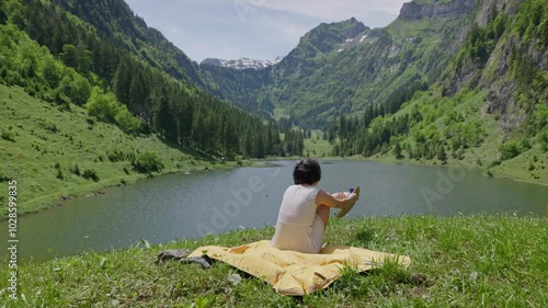 A woman stands by a mountain lake removing her hat before sitting on a blanket in the summer photo