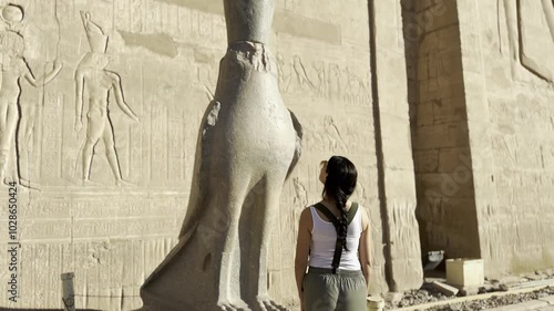 An adventurous tourist stands in awe before the majestic statue of the falcon of Horus at the temple of Edfu, experiencing a deep connection to ancient Egypt.