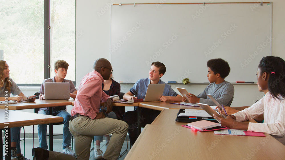 High School Teacher Helping Students Sitting At Desks Using Laptops And Digital Tablets In Classroom High School Teacher Helping Students Sitting At Desks Using Laptops And Digital Tablets In Classroom
