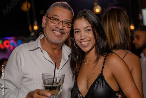 Happy young woman with her father at the club, wearing black leather dress and white shirt holding cocktail glass smiling to camera at night party